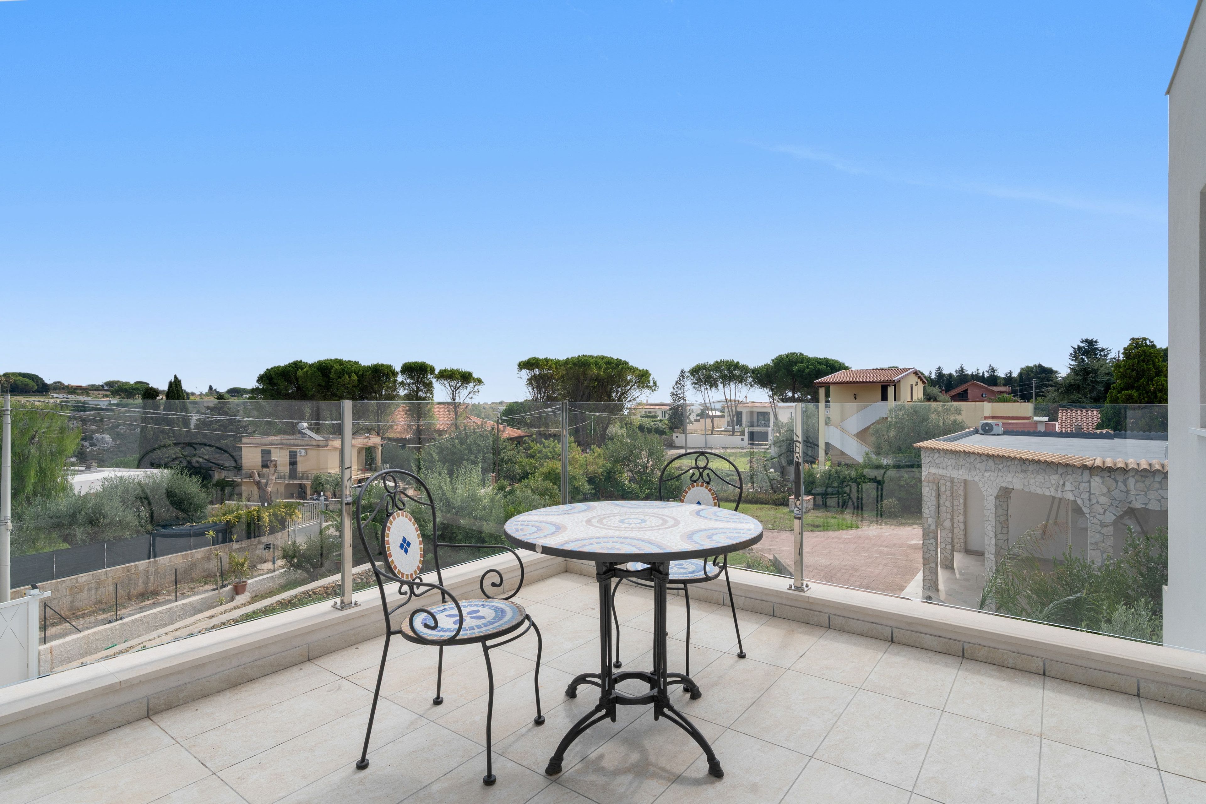 A balcony with a round table and two chairs, overlooking a residential area with houses and trees.