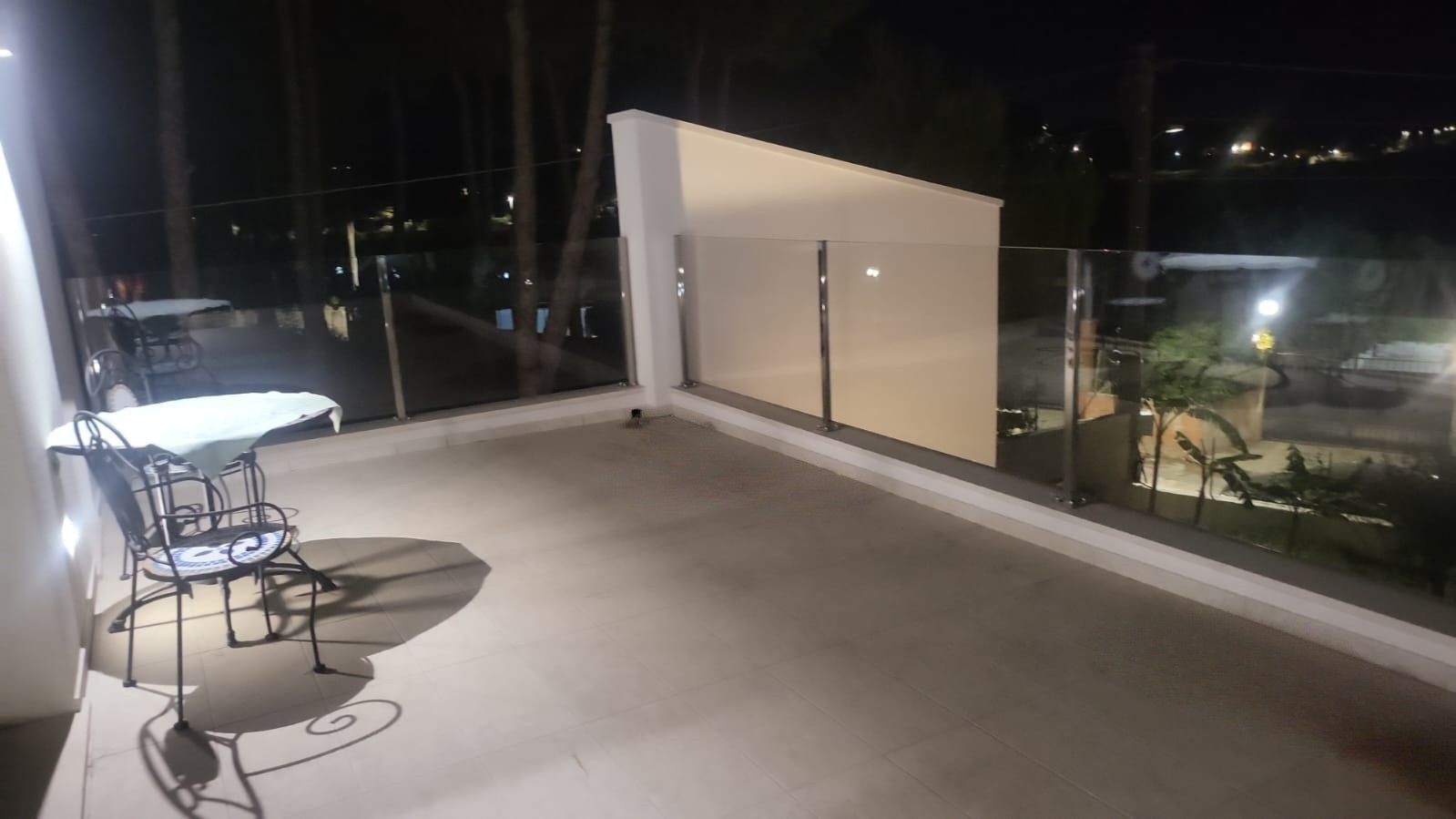 Uma varanda com mesa e cadeiras, vista da cidade à noite e guarda-corpo de vidro.