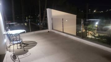 Uma varanda com mesa e cadeiras, vista da cidade à noite e guarda-corpo de vidro.