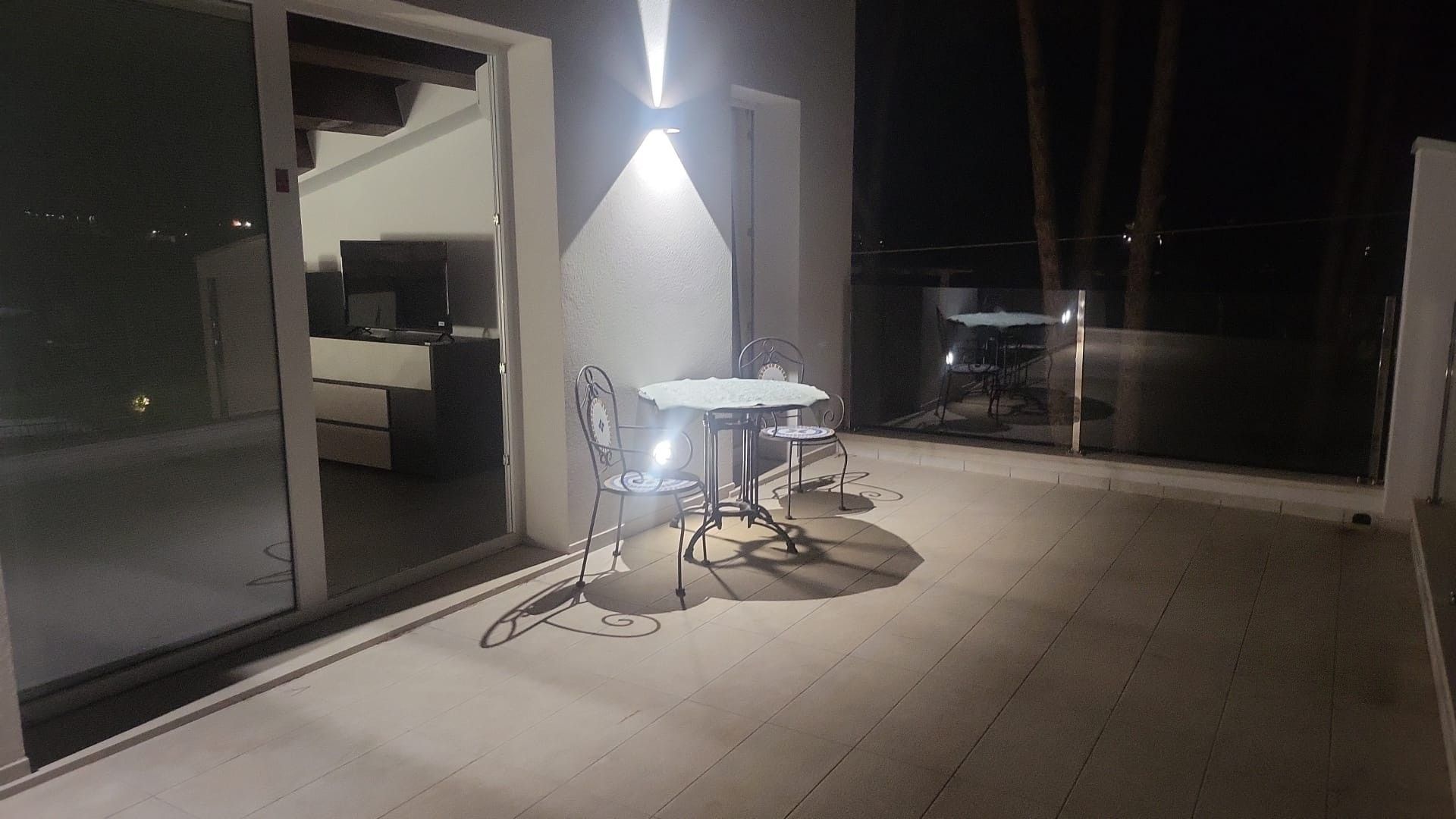 Un balcon extérieur avec une table et des chaises, une porte vitrée donnant sur une pièce et une vue sur la ville la nuit.