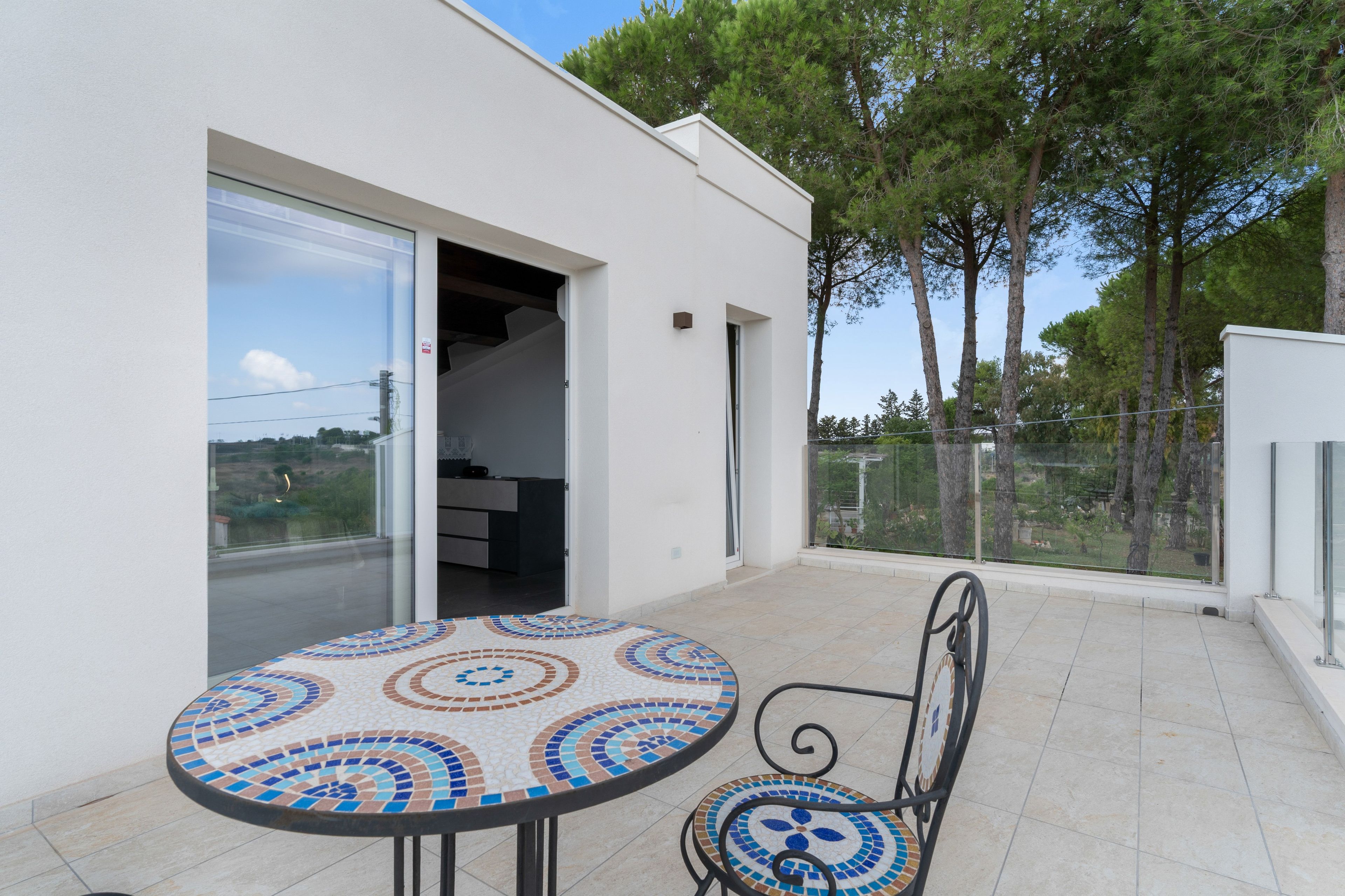 Une terrasse extérieure moderne, avec une table et des chaises carrelées, une porte-fenêtre coulissante donnant sur une pièce, et une vue sur les arbres et un ciel dégagé.