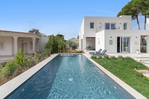 A rectangular swimming pool with a clear blue surface, surrounded by a stone wall and a grassy area with some plants.