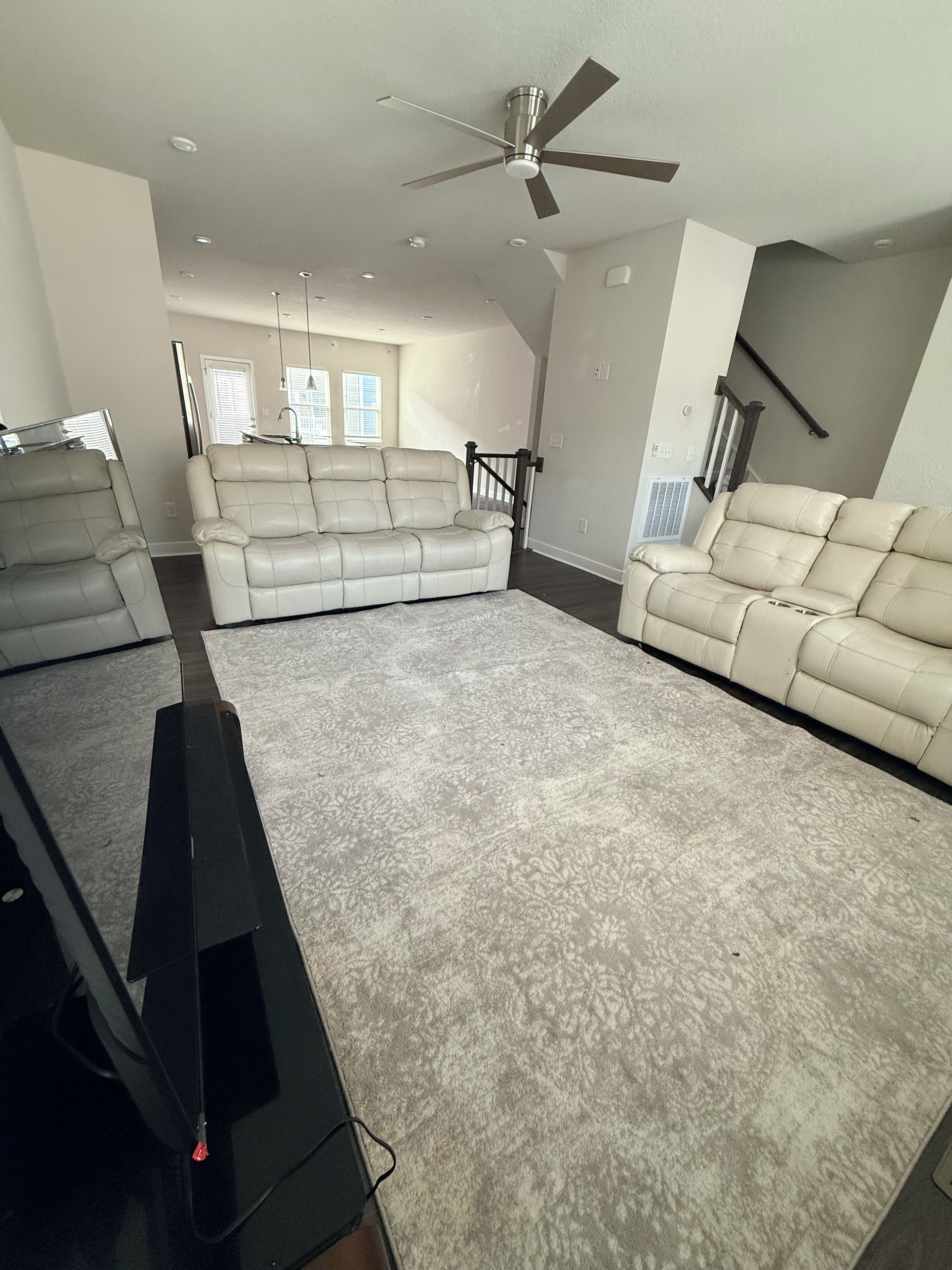 Un salón amplio con una alfombra grande color beige, dos sofás color beige, un ventilador de techo y una escalera.