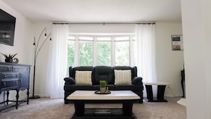 A living room with a black leather sofa, a small coffee table, and a window with white curtains. - The Summer House - Lake Access - Hot Tub & Kayaks (Lambton Shores)
