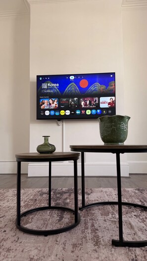 A flat-screen TV mounted on a wall displaying a travel website, a green vase on a table, and a patterned rug on the floor. - Stylish 2-Bedroom Flat in South London | Sleeps 4 (London)