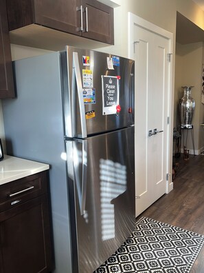 A stainless steel refrigerator with various magnets and a 'Please Clean Before You Leave' sign, a white door, and a patterned rug in a kitchen area. - 🌿 JAVA HOUSE | Private Stay, No Sharing • Free Coffee, Breakfast & Parking (Edmonton)