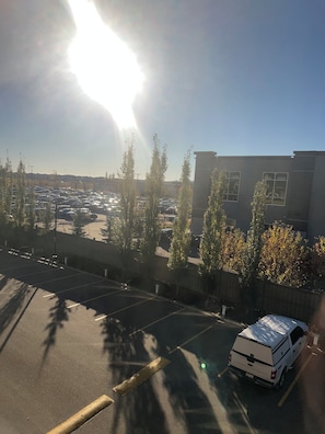 Parkkipaikka, jossa on valkoinen pakettiauto, puita ja ikkunoilla varustettu rakennus.