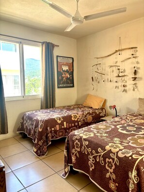 A room with two beds, a ceiling fan, a poster on the wall, and a window with curtains. - Excellent location (Puerto Vallarta)