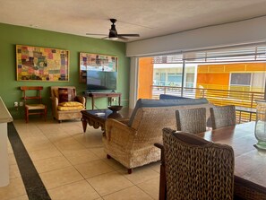 A living room with a green wall, a ceiling fan, a television, and a dining area with wicker furniture. - Excellent location (Puerto Vallarta)