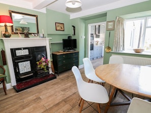 A dining area with a wooden table and white chairs, a fireplace with a red mantle, and a kitchen visible through an open door. - Ivy Cottage (Buxton)
