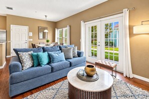 A living room with a blue sofa, a round coffee table, and a view of a green lawn through a sliding glass door. - 15mins Disney~Themed Suites~Resort Pool~Game Room (Kissimmee)
