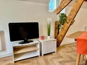 Ein modernes Wohnzimmer mit einem Flachbildfernseher auf einem weißen Schrank, einem kleinen weißen Tisch mit einem rosa Kerzenlicht, einer Topfpflanze und einer Holztreppe.