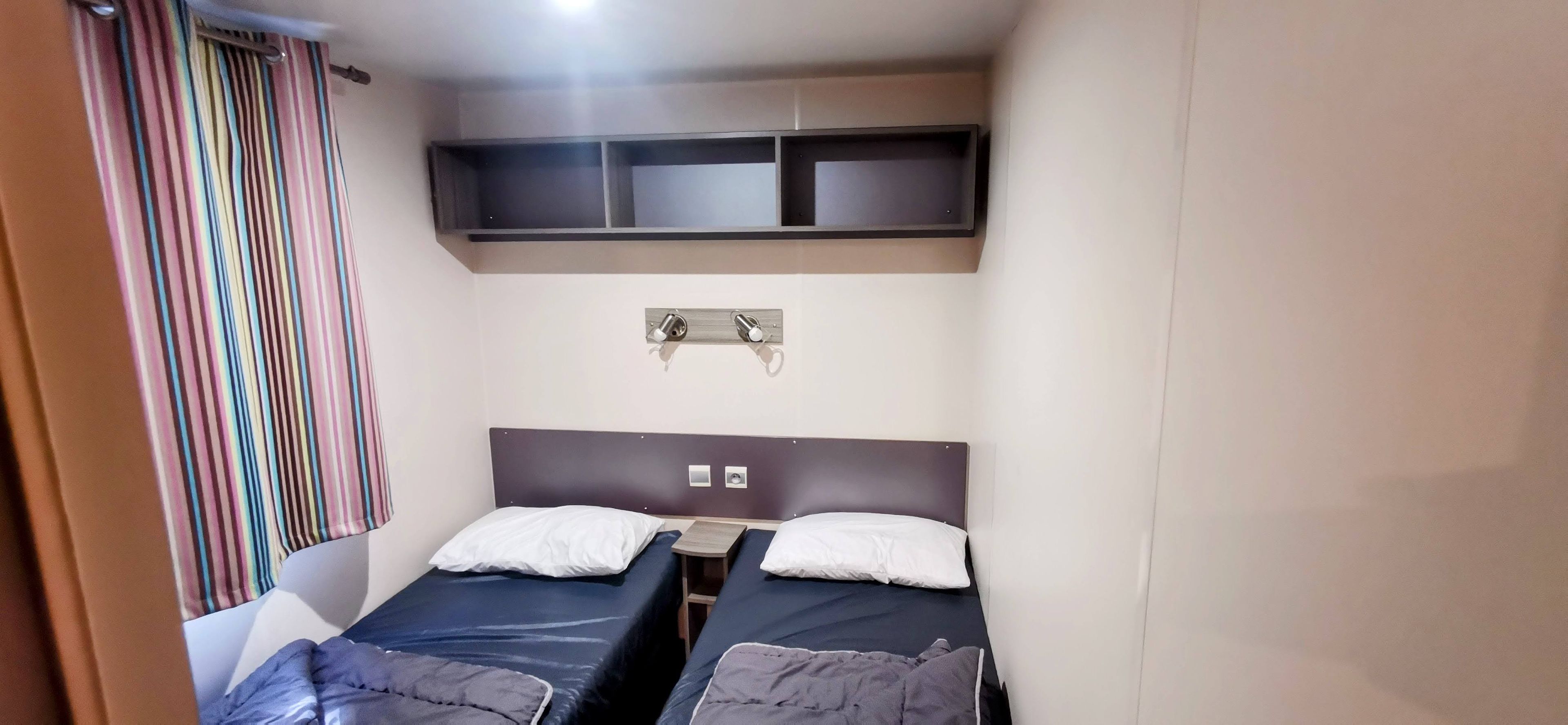 Quarto pequeno com duas camas de solteiro, cortina listrada e prateleira sobre as camas.