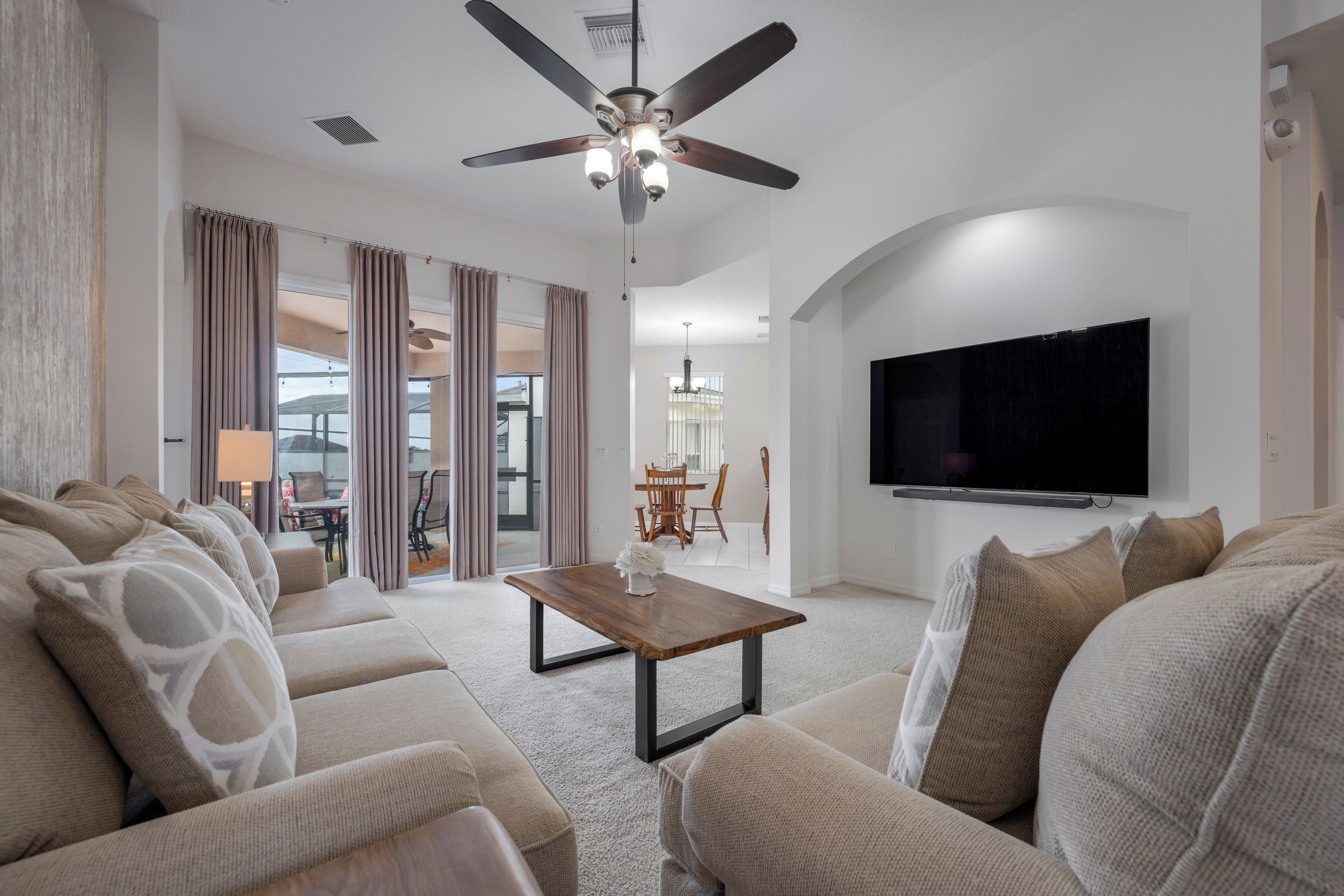 A modern living room with a ceiling fan, a flat-screen TV mounted on the wall, and a dining area visible through a glass door.