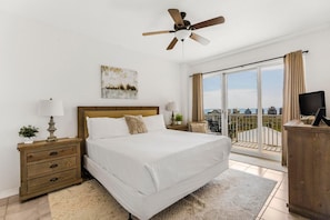 Un dormitorio con cama, mesita de noche, lámpara, ventilador de techo, ventana con cortinas y vistas al exterior.