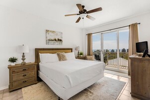 Un dormitorio con cama, mesita de noche, lámpara, ventilador de techo, ventana con cortinas y vistas al exterior.