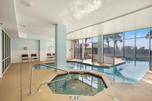 Piscina interior con jacuzzi, paredes de vidrio transparente y vistas a palmeras.