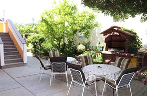Eine Terrass mit einem Backstein-Barbecue, weißen Metallstühlen und einer Treppe zu einer erhöhten Ebene.