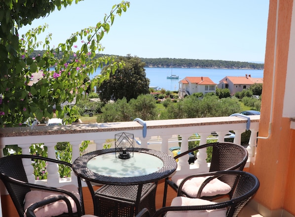 Ein Balkon mit Korbmöbeln und einem Glastisch, der auf eine Küstenlandschaft mit Häusern und Booten blickt.