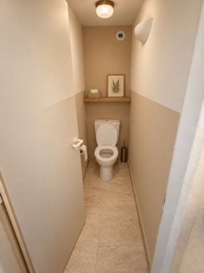 Un baño pequeño con inodoro, repisa con una hoja enmarcada y dispensador de papel higiénico.