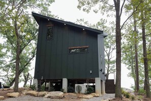 A cabin with a dark exterior, a small window, and a staircase leading up to the entrance. - The Ranger - Luxury Treehouse (Pikeville)