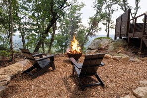 A fire pit with two Adirondack chairs in a forested area with a view of the water. - The Ranger - Luxury Treehouse (Pikeville)
