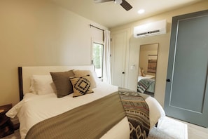 Ein Schlafzimmer mit einem Bett, Kissen, einer Decke, einem Deckenventilator, einer Klimaanlage, einer Tür und einem Fenster.