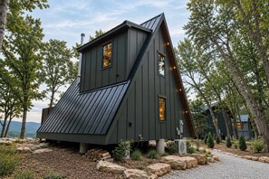 Eine moderne, dunkel gefärbte Holzhütte mit geneigtem Dach, umgeben von Bäumen und einem Kiesweg.