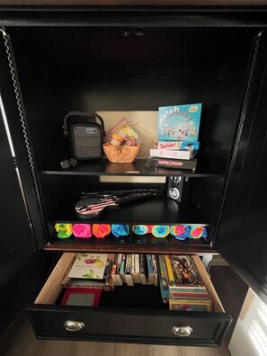 A black storage unit with shelves containing books, games, and a small bag. - Calm, cozy home-large patio w/ mountain view. (Mattawa)