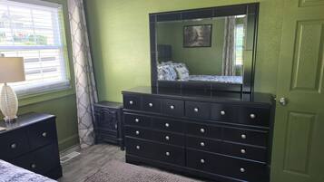 Une chambre à coucher avec une commode, un lit, une fenêtre avec des stores et une porte.