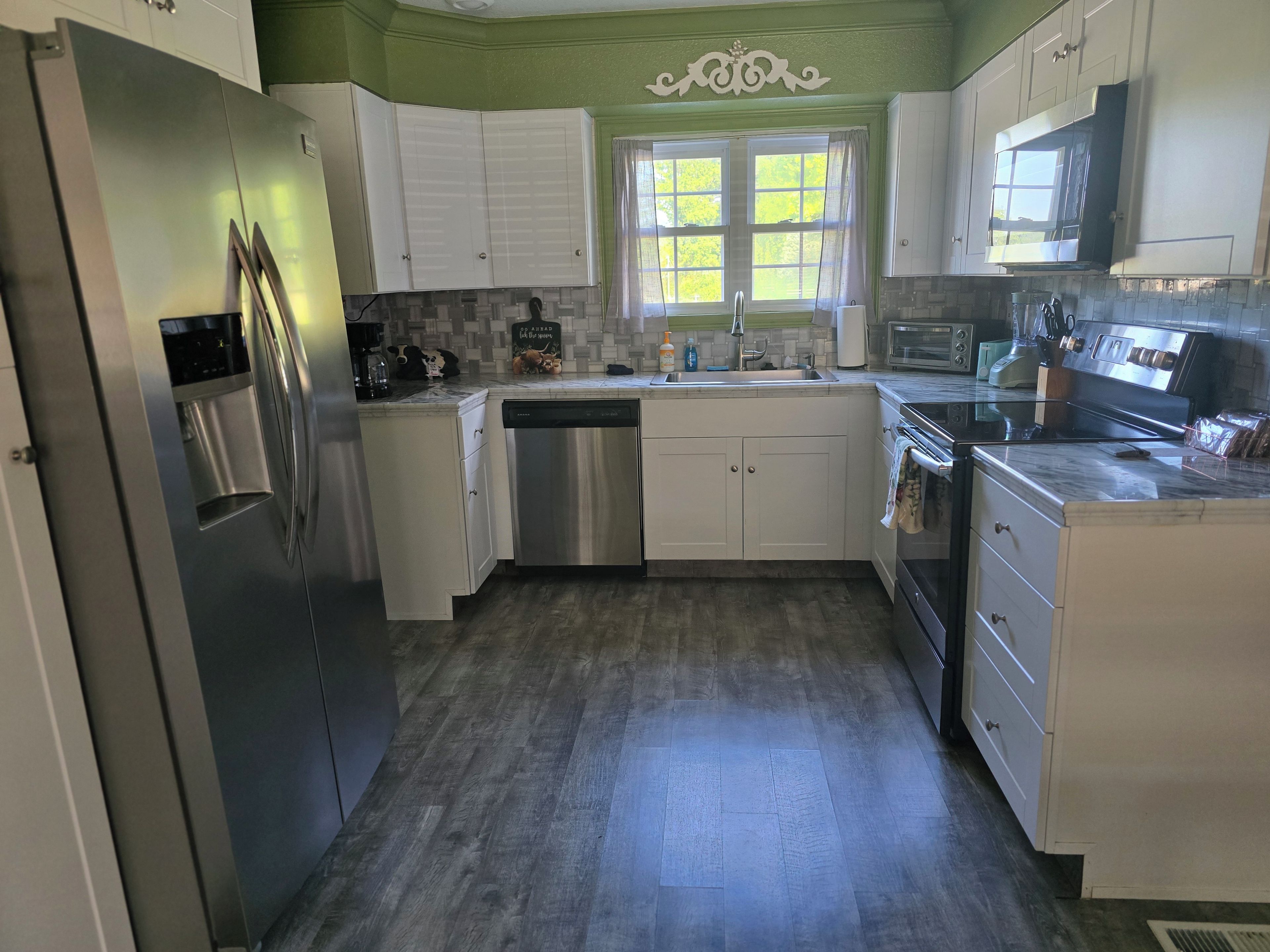 Cozinha com armários brancos, eletrodomésticos de aço inoxidável, janela com cortinas e painel de azulejos.