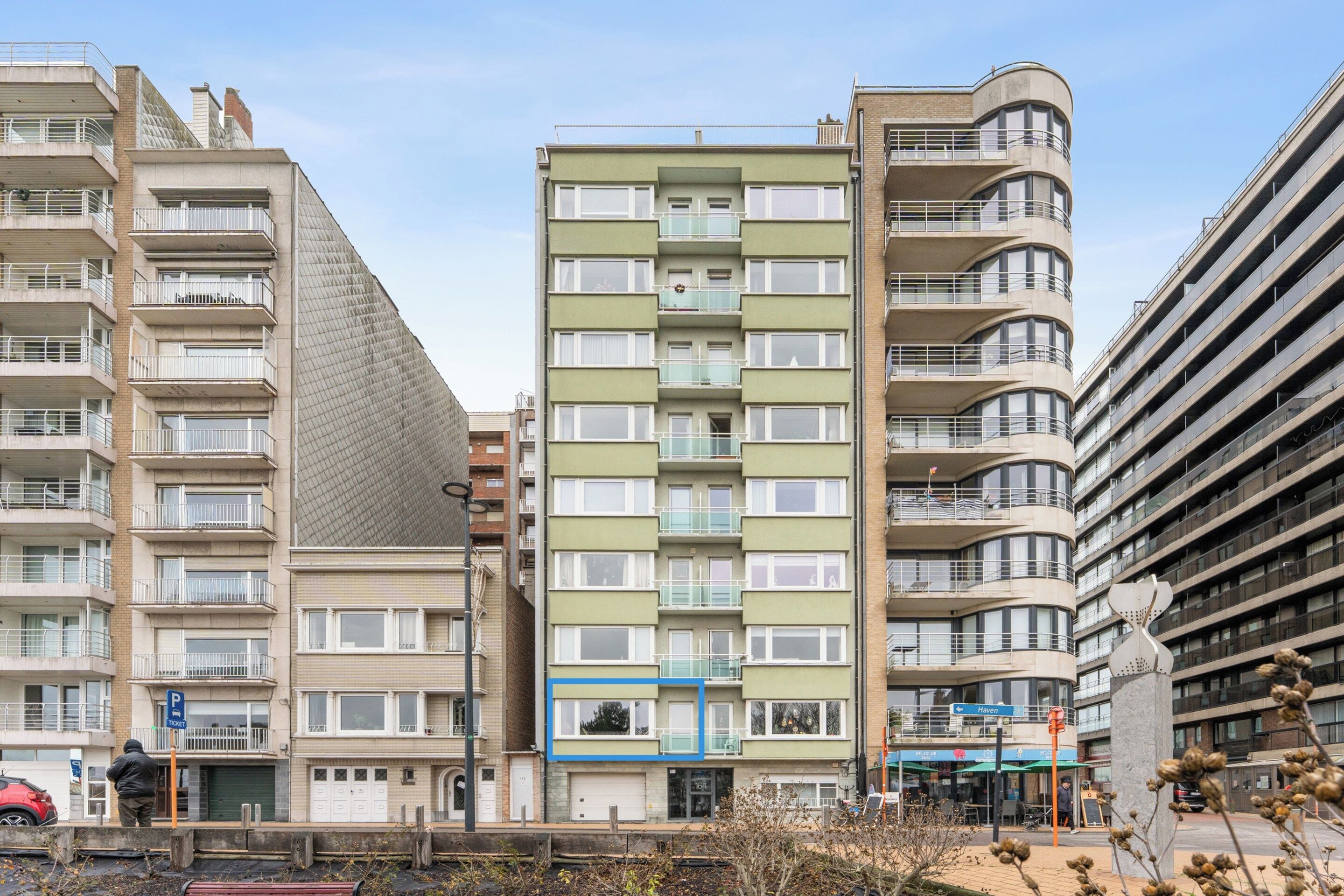 A multi-story residential building with balconies, a parking area, and a street view.