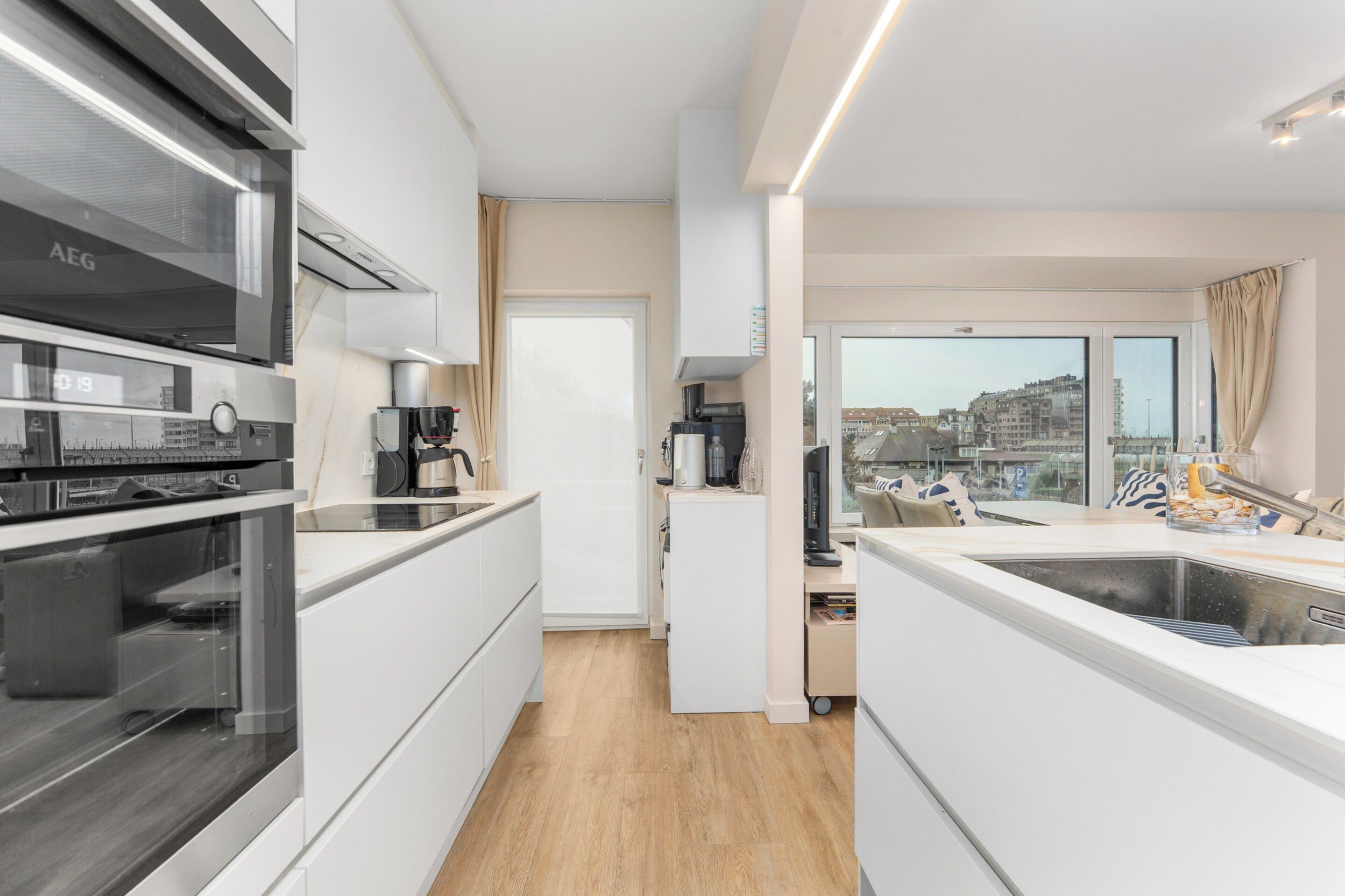 A modern kitchen with a built-in oven, a coffee machine, and a view of the city through large windows.