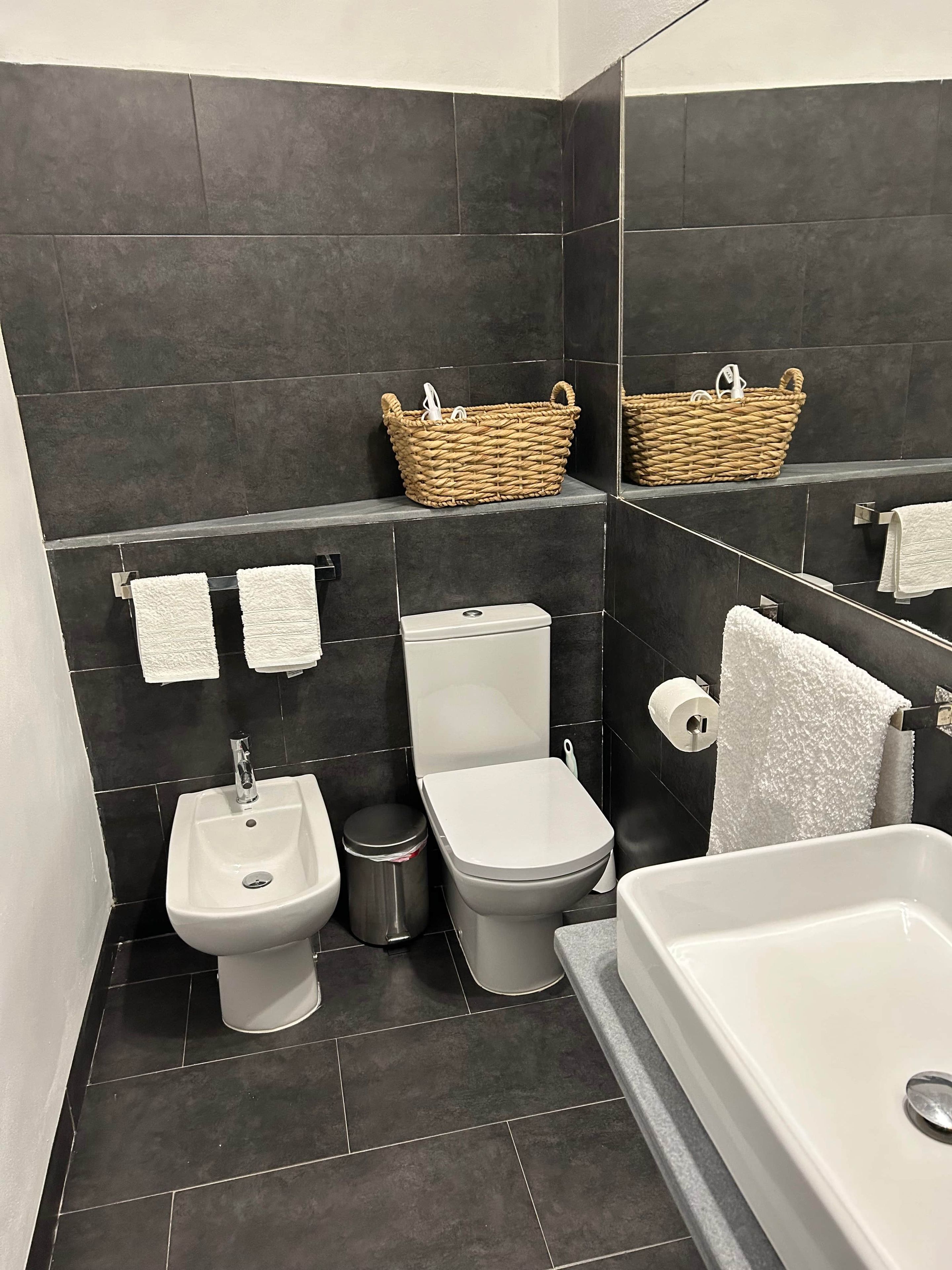 Une salle de bain moderne équipée d’un bidet, de toilettes et d’une baignoire. On y trouve deux paniers en osier accrochés au mur et une poubelle.