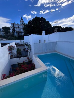 Une piscine à l’extérieur blanc, entourée d’un mur blanc et d’une clôture. On y trouve un balcon agrémenté de plantes et de fleurs en pot.