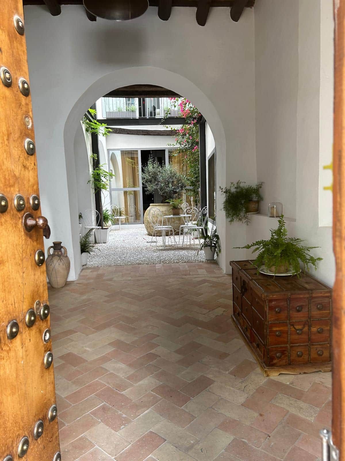 Un couloir intérieur avec une porte en bois, un sol carrelé et une vue sur une pièce dotée d’une grande fenêtre, d’un meuble en bois et de plantes en pot.