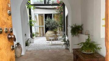 Un couloir intérieur avec une porte en bois, un sol carrelé et une vue sur une pièce dotée d’une grande fenêtre, d’un meuble en bois et de plantes en pot.