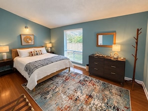 A bedroom with a bed, bedside lamps, a dresser, a mirror, and a window with blinds. - Palm & Pine  (Ocean Springs)