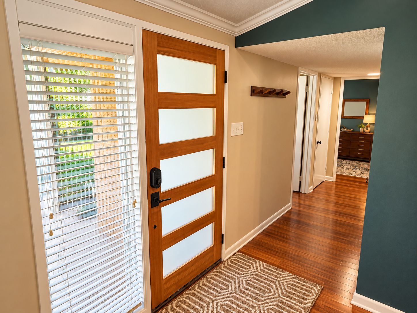 Un pasillo con una puerta de madera, un tapete estampado y vista a una habitación con cómoda y l ámpara.