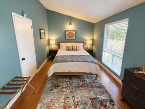Un dormitorio con una cama, lámparas de noche, una alfombra estampada, un aparador y una ventana con persianas.