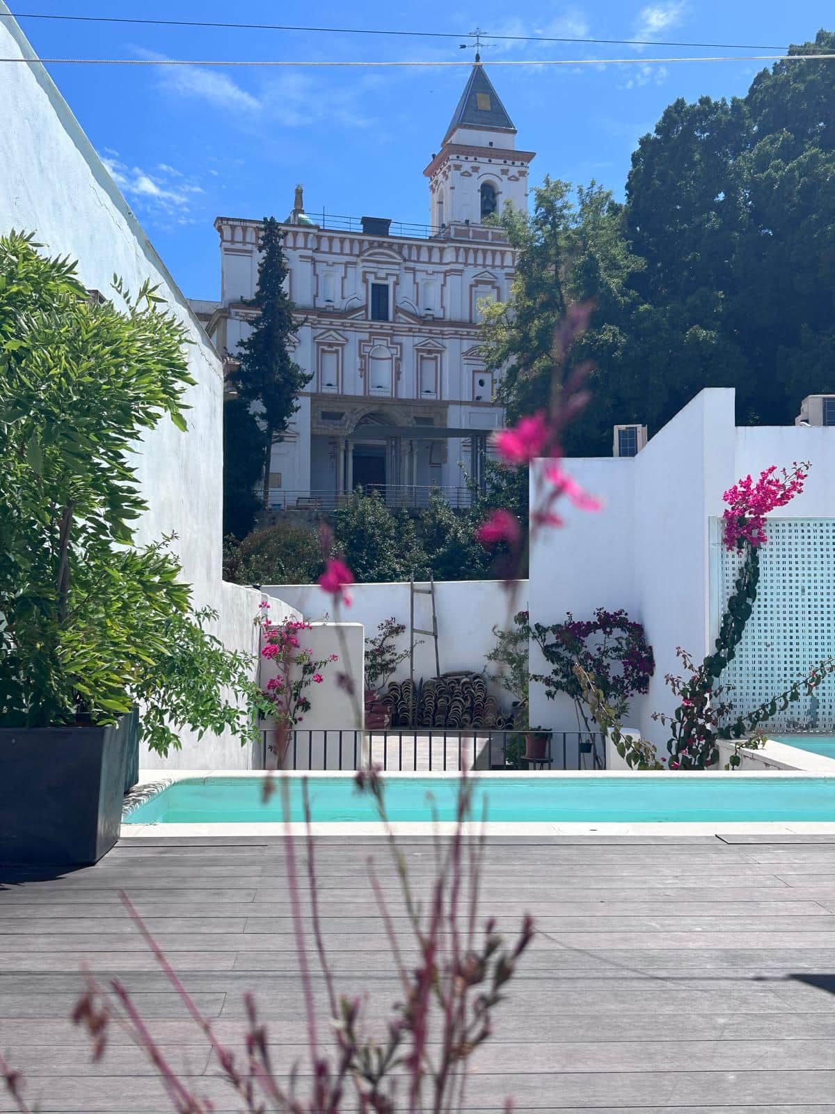 A courtyard with a pool, a white building with a tower, and surrounding greenery.
