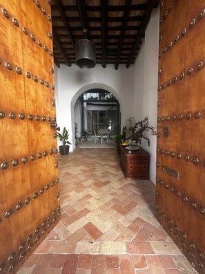 Un couloir au sol orné d’un motif en chevrons, bordé de portes en bois rehaussées de clous en métal, qui mène à une porte en arc de cercle.