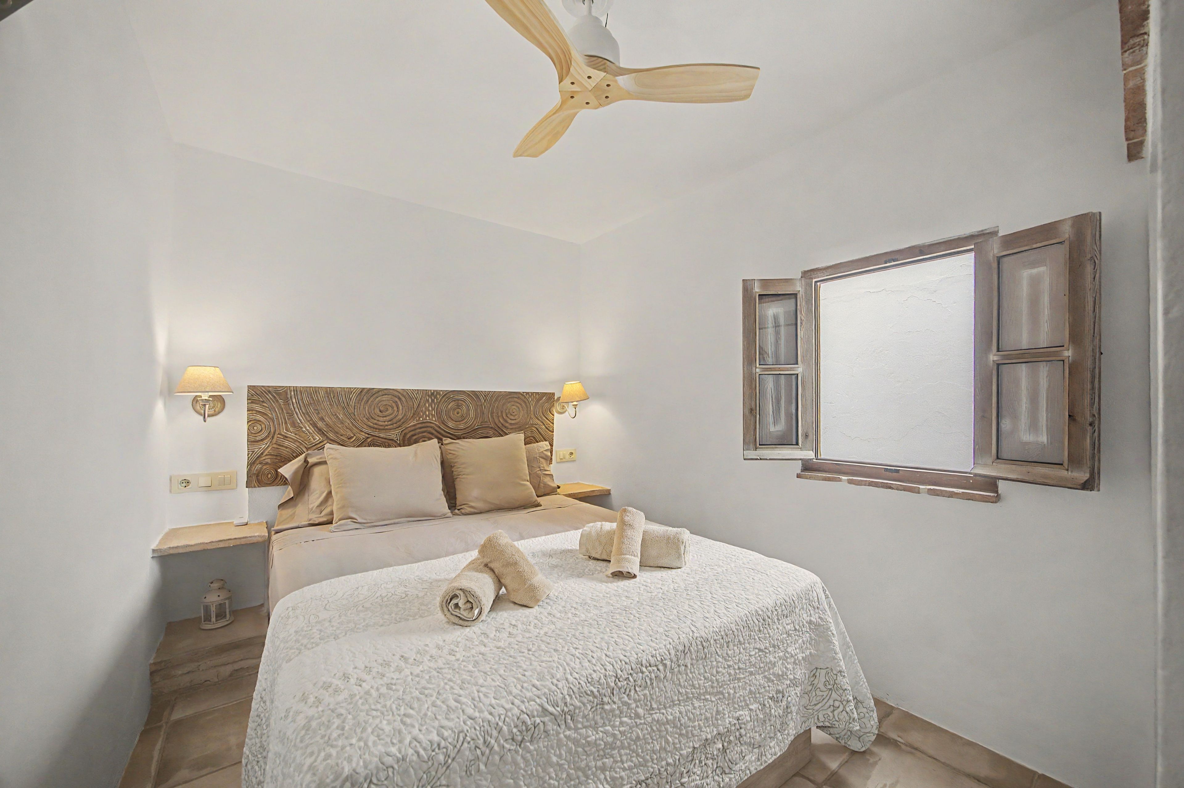A bedroom with a bed, a ceiling fan, bedside lamps, a window with shutters, and a nightstand.