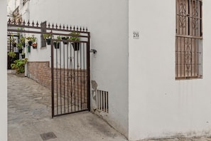 Una entrada protegida por una reja a un edificio con fachada blanca, sección inferior de ladrillo y una ventana con rejas de hierro decorativas.