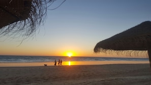 Escena de playa con atardecer, gente caminando y una estructura de techo de paja.
