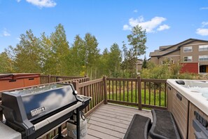 A deck with a barbecue grill, hot tub, and wooden railing overlooking a landscaped area with trees and a building in the background. - New 2-bedroom townhome with private hot tub (Silverthorne)