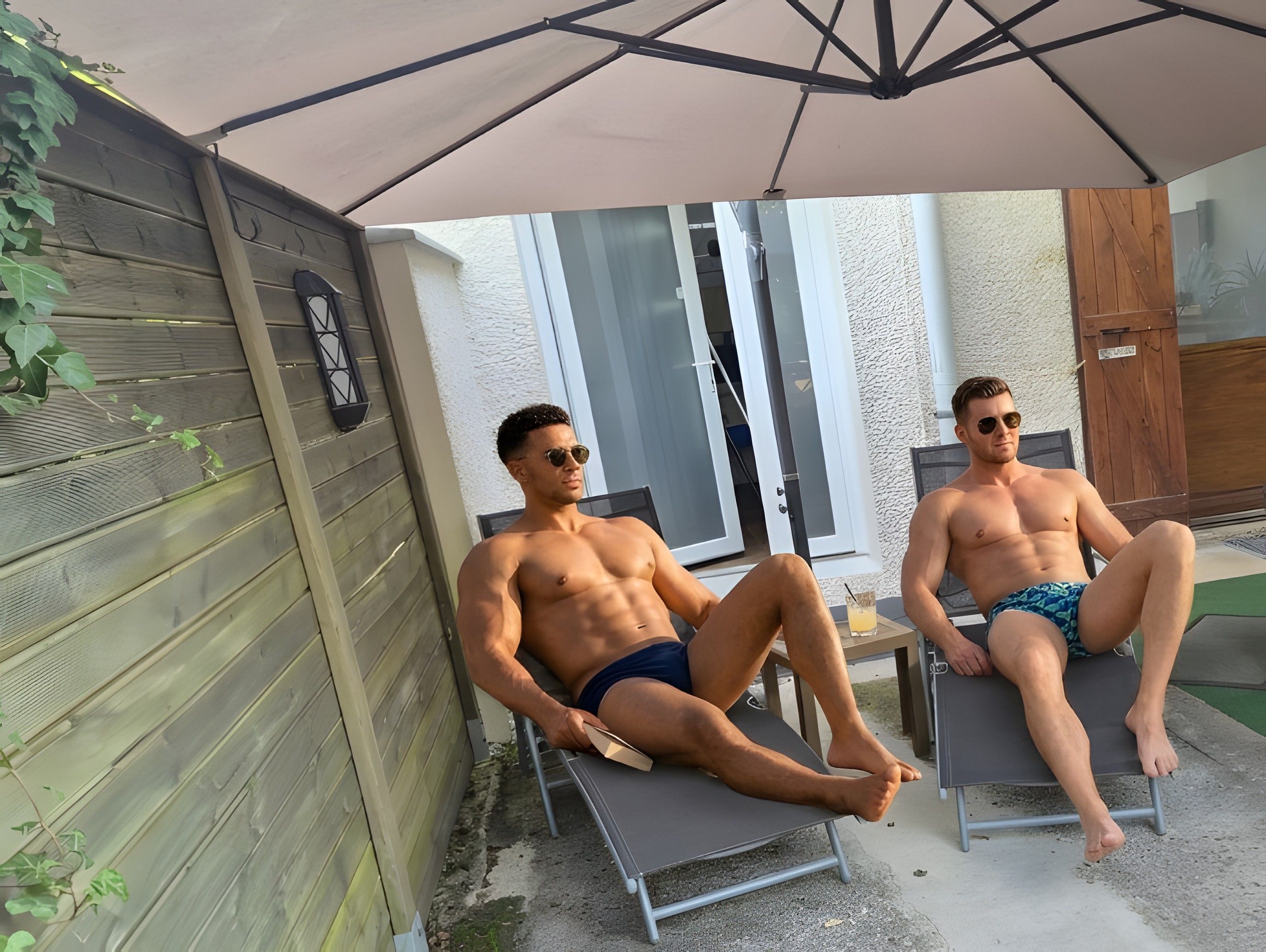 Deux hommes sont assis sur des chaises longues, à l’ombre d’un parasol, dans un espace extérieur aménagé en patio.