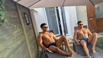 Deux hommes sont assis sur des chaises longues, à l’ombre d’un parasol, dans un espace extérieur aménagé en patio.