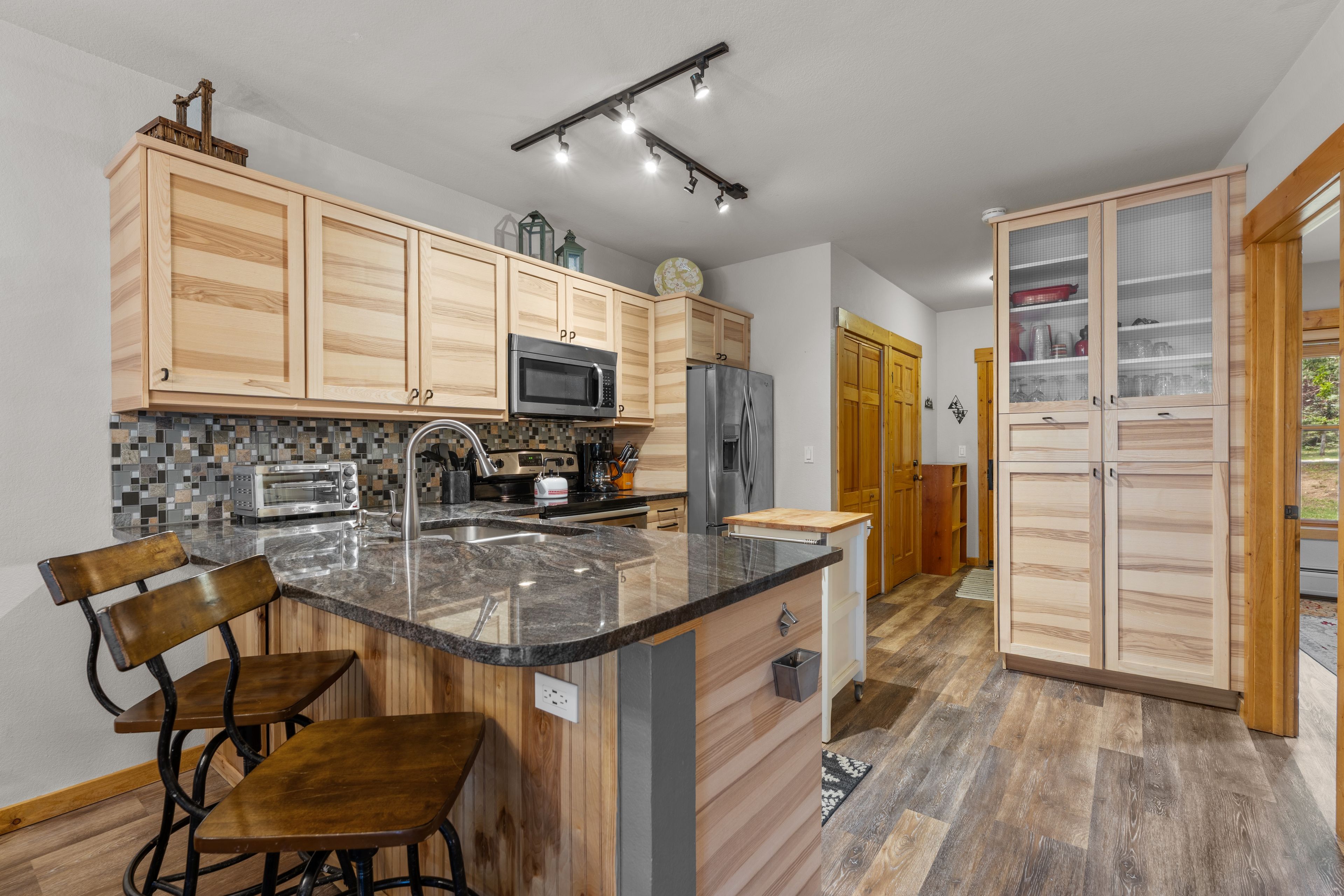 A modern kitchen with wooden cabinets, a central island, bar stools, and a stainless steel refrigerator.