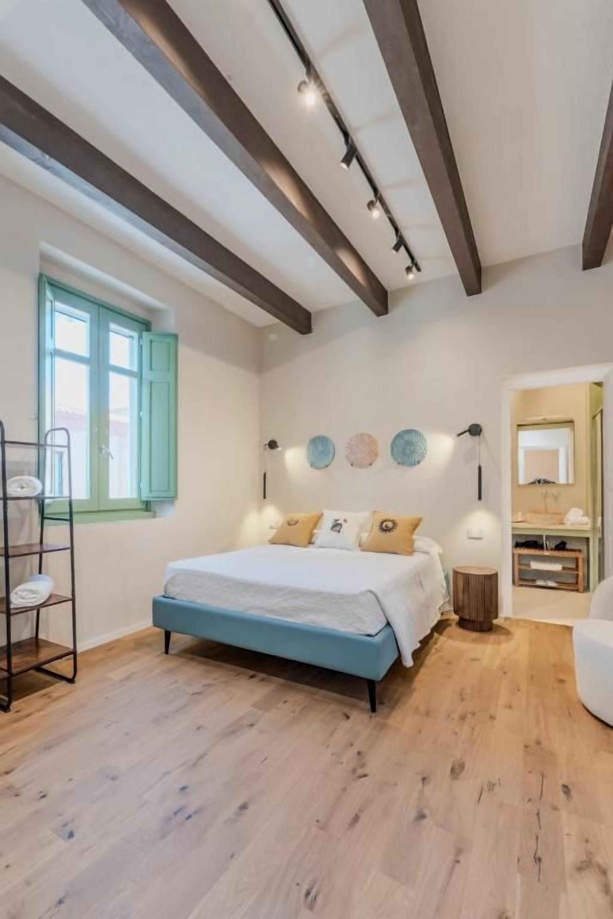 Un dormitorio con cama, piso de madera, ventana y mesita de noche.